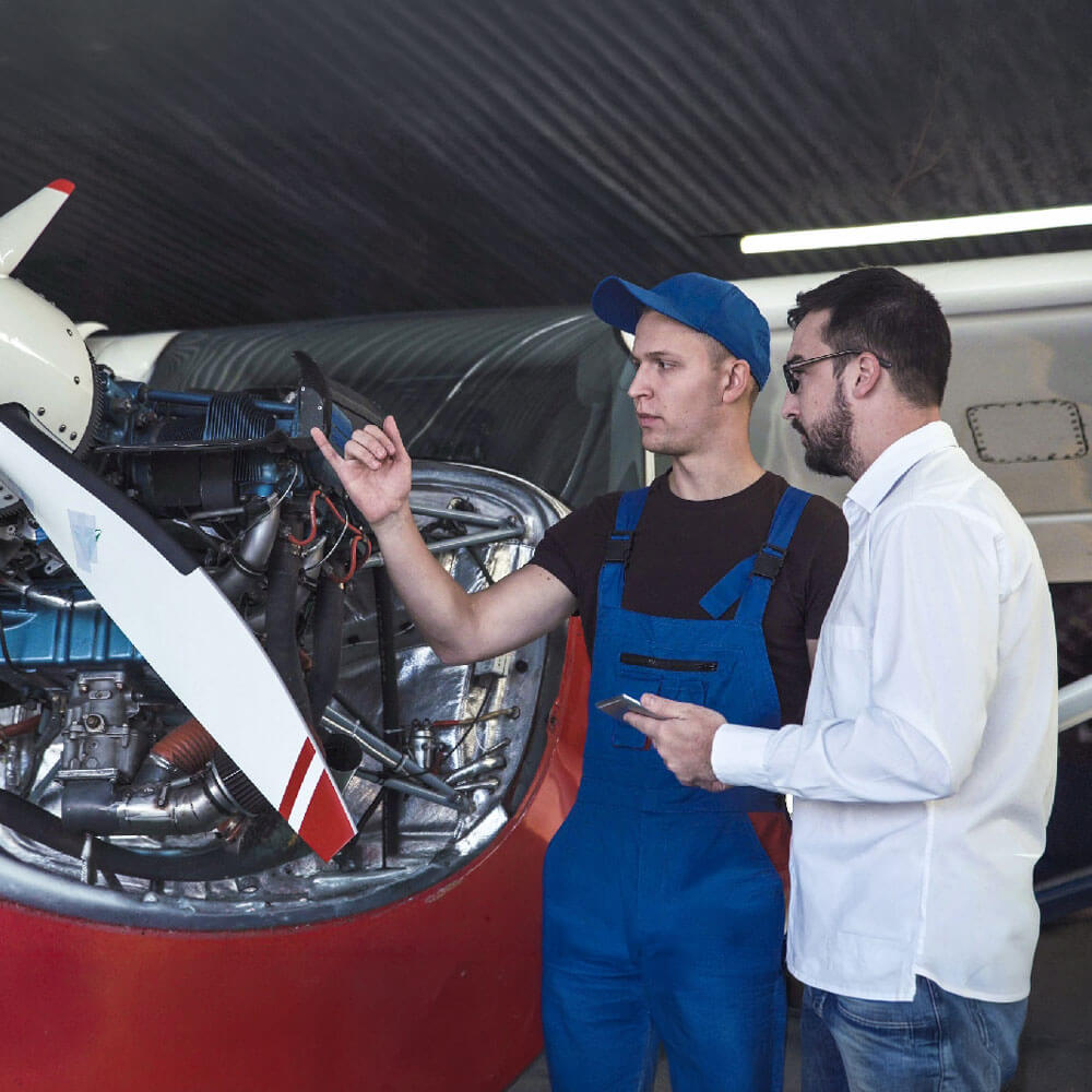 A mechanic points to an aircraft engine while explaining something to another man in a hangar.