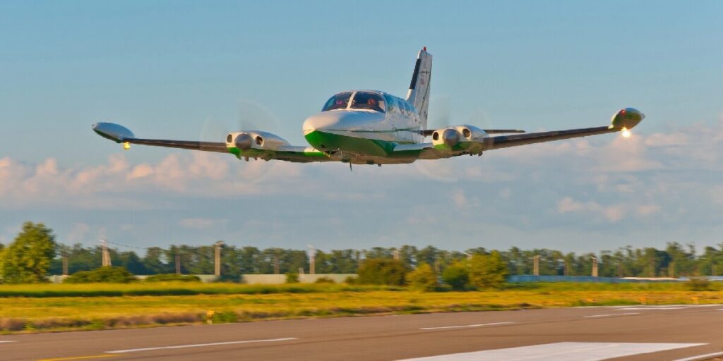 Twin engine propeller plane taking off low over a runway against a blue sky.