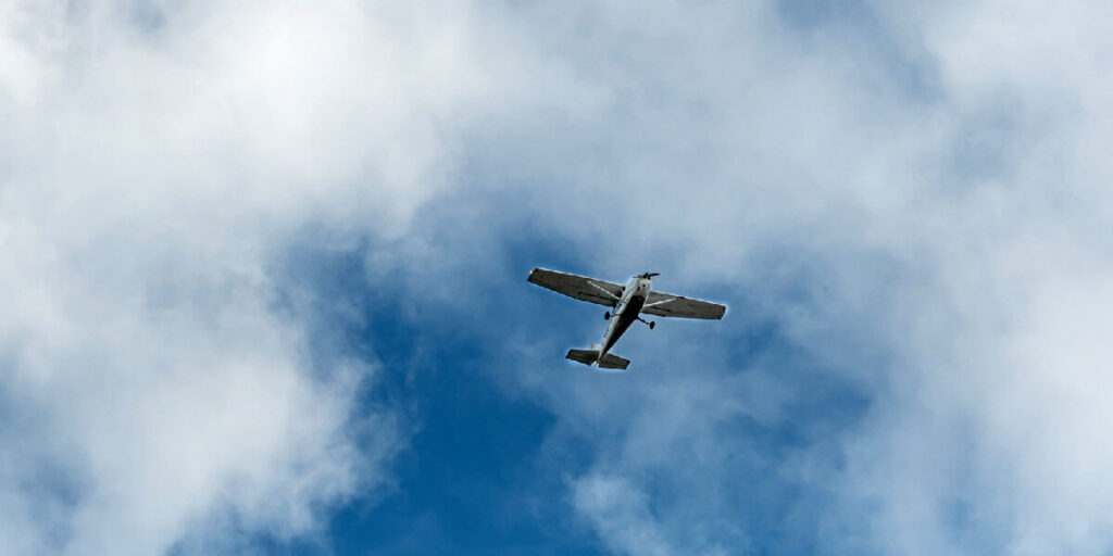 A small single-engine plane flies through a bright blue sky dotted with clouds.