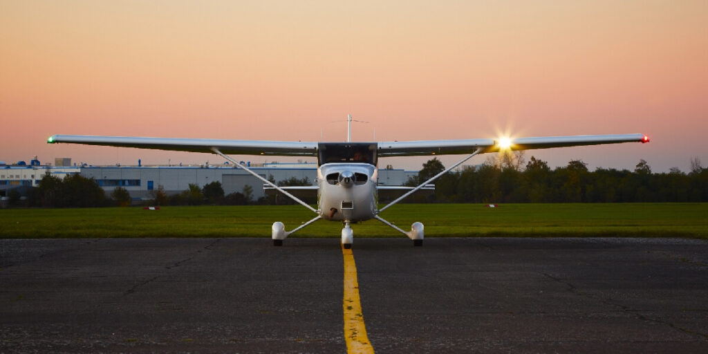 A small single-engine plane faces forward on a taxiway at sunset with its wing light glowing.